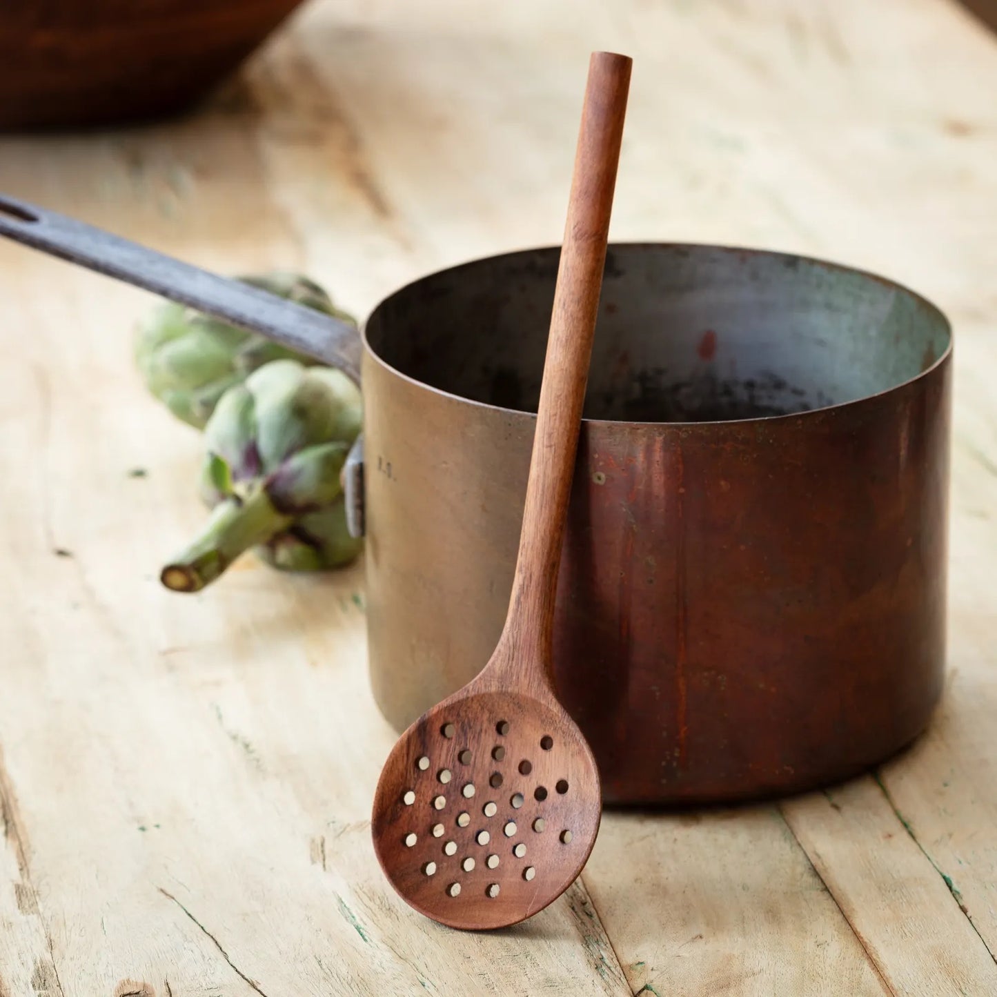 Rustic wooden straining spoon with perforations, leaning against a vintage pot. A functional and display-worthy kitchen tool for cooking and serving.