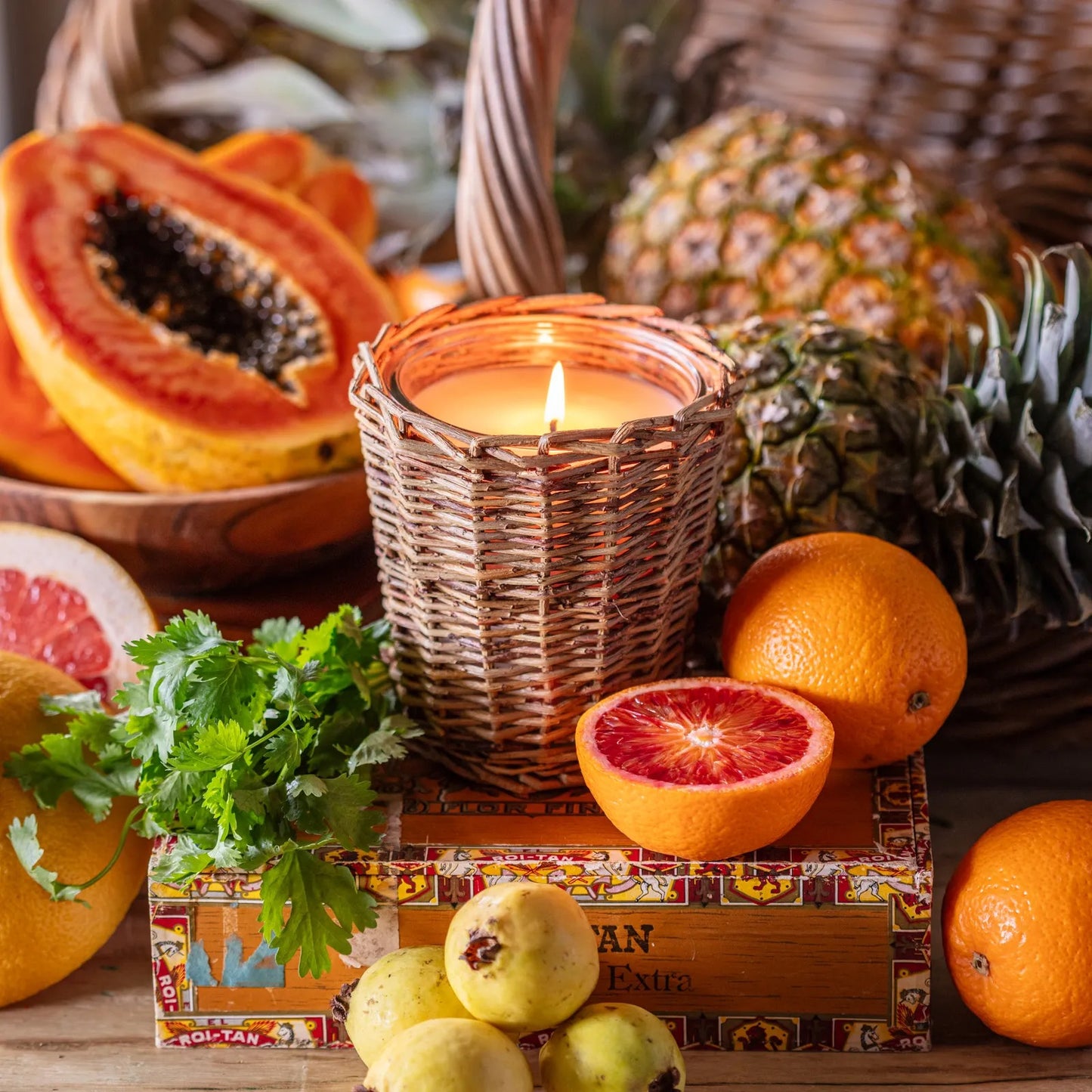 Porch View Home - Havana Candle in a woven holder, surrounded by fresh pineapple, papaya, oranges, and herbs. Features a lemon pineapple scent.