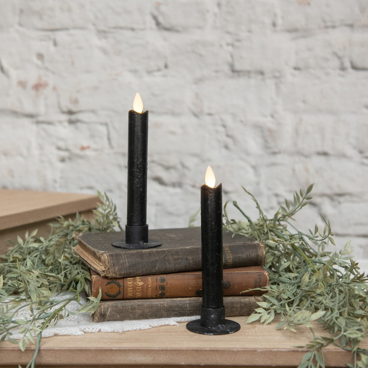 Two 6.75-inch black taper candles with moving flame effect displayed on vintage books with greenery. Elegant flameless candles for cozy decor.