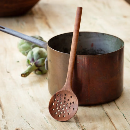 Rustic wooden straining spoon with perforations, leaning against a vintage pot. A functional and display-worthy kitchen tool for cooking and serving.