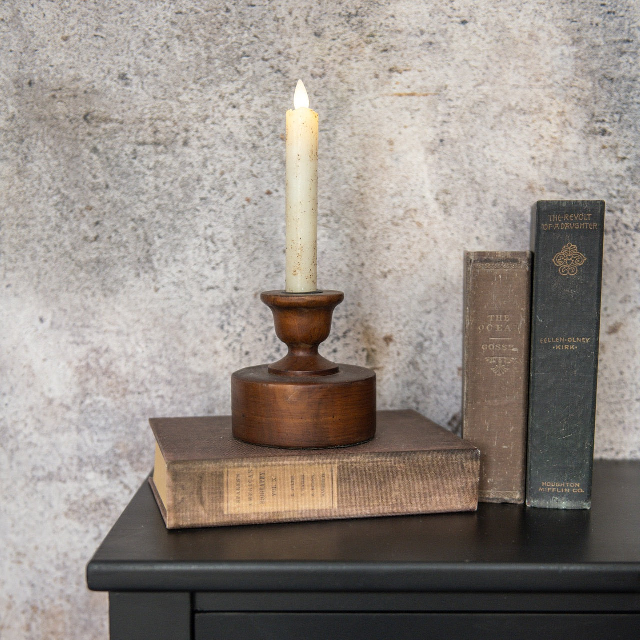 MENTOR TAPER CANDLESTICK with a lit candle, placed on stacked vintage books atop a black wooden table. The handcrafted wooden holder adds rustic charm to the decor.