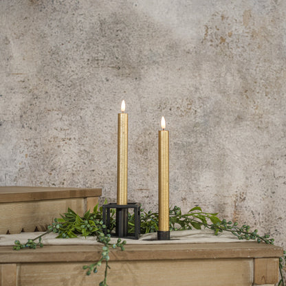 Set of two 9.5-inch 3D Flame Gold Taper Candles with a realistic flickering glow, displayed on a rustic wooden surface with greenery accents.