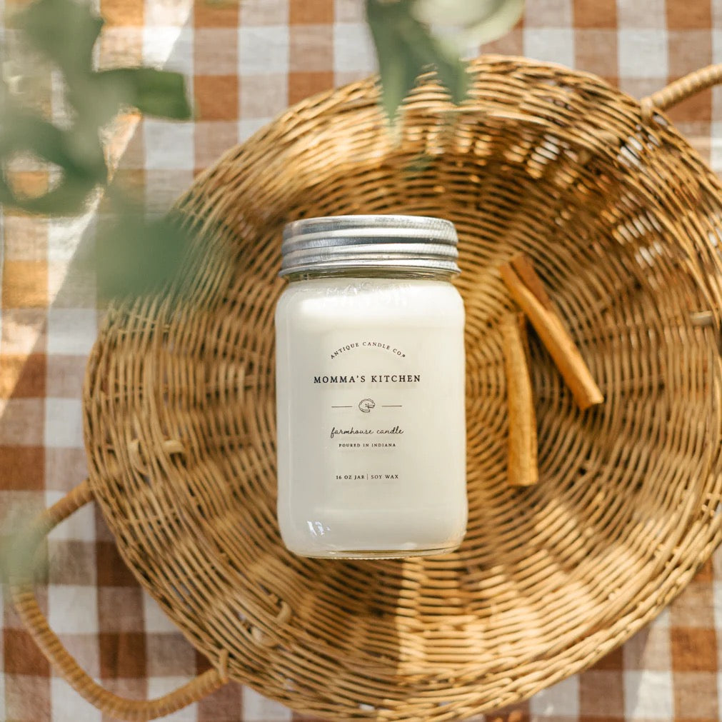 Antique Candle Co 16oz soy candle in 'Momma's Kitchen' scent, placed on a woven basket with cinnamon sticks. Long-lasting home fragrance with natural soy wax.