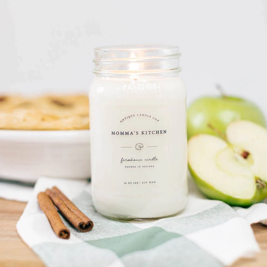 Antique Candle Co 16oz 'Momma's Kitchen' soy candle burning beside fresh apples and cinnamon sticks. Cozy farmhouse scent with long burn time.