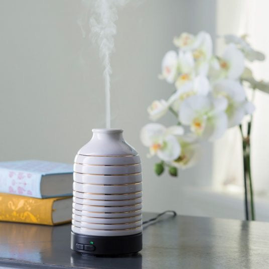 Serenity Ultra Sonic Diffuser emitting a fine mist, placed on a table with books and white orchids, enhancing home ambiance with aromatherapy.