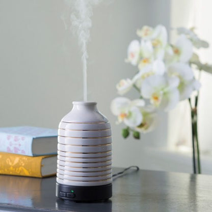 Serenity Ultra Sonic Diffuser emitting a fine mist, placed on a table with books and white orchids, enhancing home ambiance with aromatherapy.