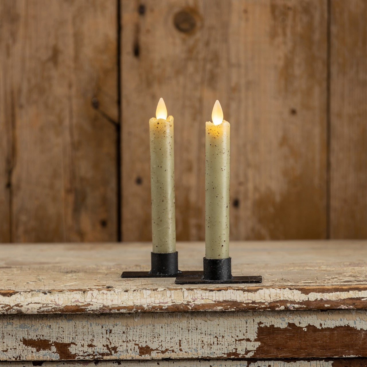 Set of two 6.75-inch moving flame green taper candles with a rustic finish, placed on black metal holders against a wooden backdrop.