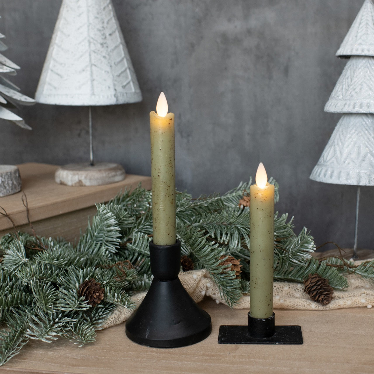 Festive display of moving flame green taper candles with holiday decorations, including pine branches and white Christmas trees.