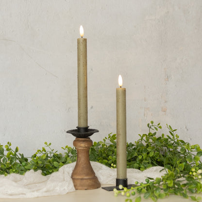 Set of two 9.5-inch green taper candles with realistic 3D flame, displayed in black and wooden candle holders with greenery decor.
