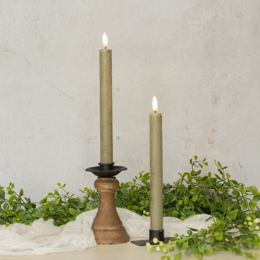 Set of two 9.5-inch green taper candles with realistic 3D flame, displayed in black and wooden candle holders with greenery decor.
