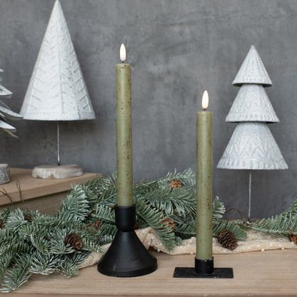 Pair of 9.5-inch green taper candles with 3D flame, placed in black holders among winter-themed decor with white Christmas trees and pine branches.