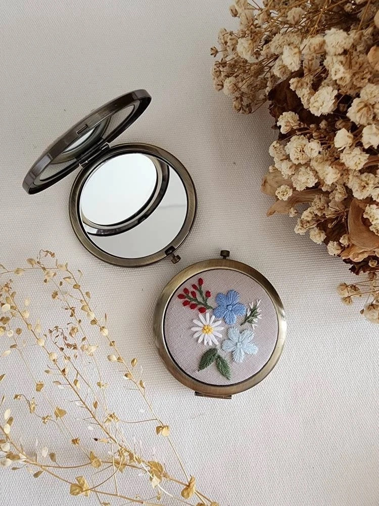Open floral embroidered compact mirror from Quince Fables, featuring a delicate blue and white flower design on a beige fabric cover.