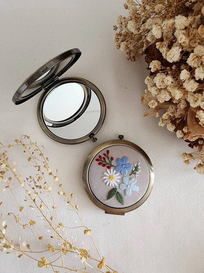 Open floral embroidered compact mirror from Quince Fables, featuring a delicate blue and white flower design on a beige fabric cover.