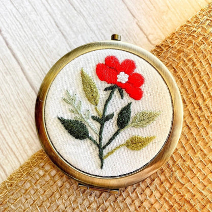 Handcrafted floral embroidered compact mirror from Quince Fables, featuring a red flower on a cream fabric background.