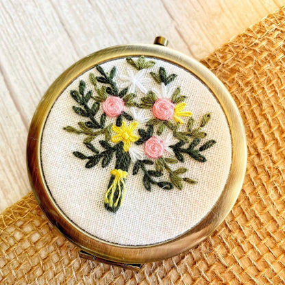 Floral embroidered compact mirror from Quince Fables, showcasing a bouquet of pink and yellow flowers on a cream fabric cover.