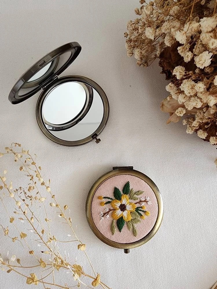 Floral embroidered compact mirror from Quince Fables, featuring a yellow flower design on a soft pink fabric cover.