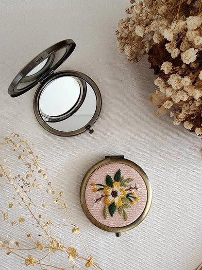 Floral embroidered compact mirror from Quince Fables, featuring a yellow flower design on a soft pink fabric cover.