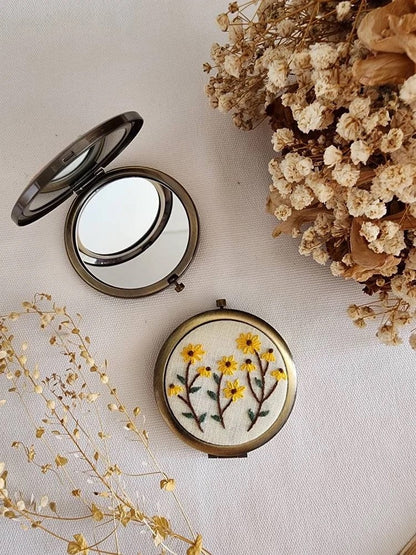 Floral embroidered compact mirror from Quince Fables, showcasing yellow flowers on a cream fabric background.