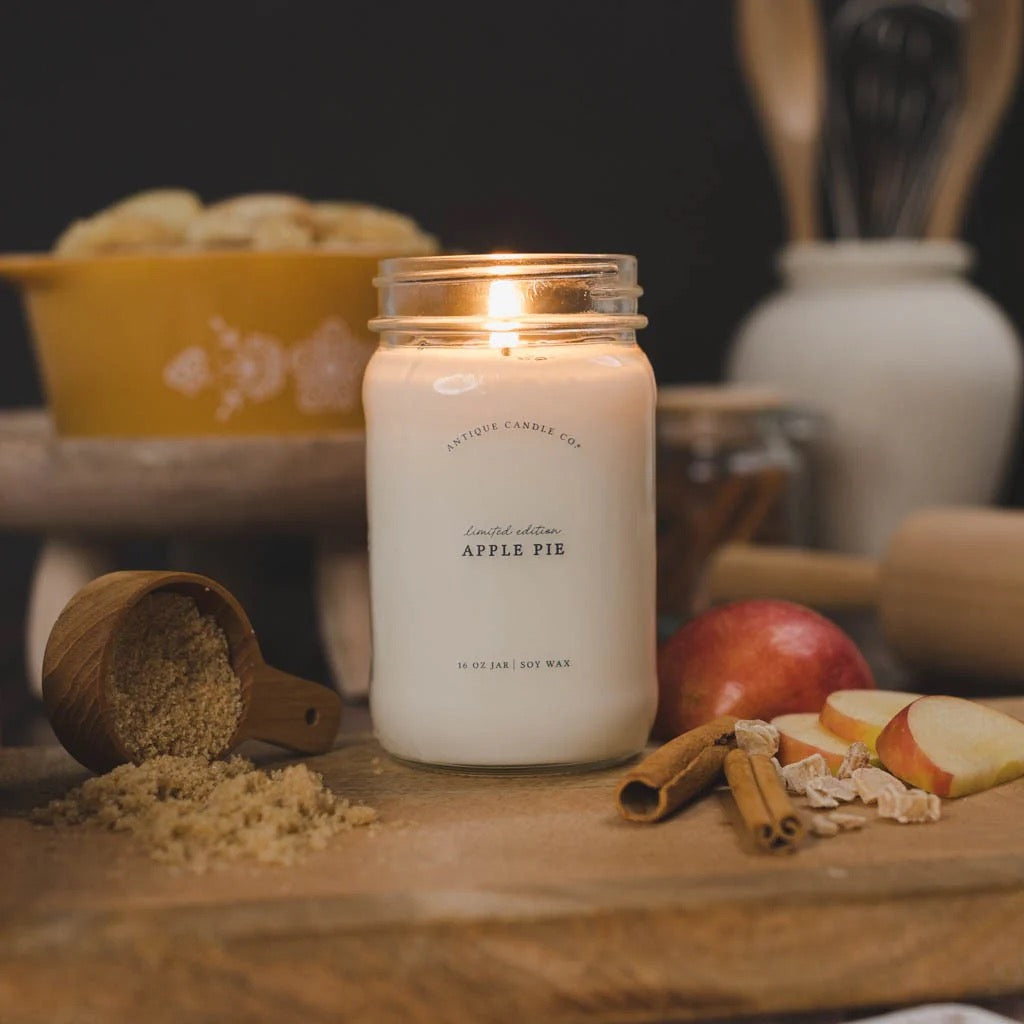 Antique Candle Co 16oz 'Apple Pie' soy candle lit on a rustic kitchen counter with brown sugar and cinnamon. Warm, inviting home fragrance.
