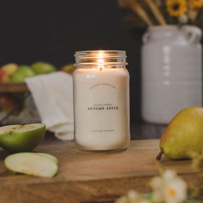 Antique Candle Co 16oz 'Autumn Apple' soy candle lit on a wooden surface with apples and pears. Cozy seasonal scent with long burn time.