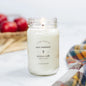 Antique Candle Co 16oz 'Fall Harvest' soy candle burning beside red apples and a plaid cloth. Warm autumn fragrance with natural soy wax.