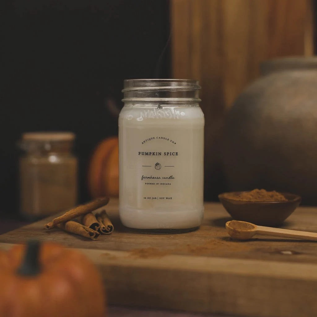 Antique Candle Co 16oz 'Pumpkin Spice' soy candle placed on a rustic wooden table with cinnamon and pumpkin. Cozy fall scent with long burn time.