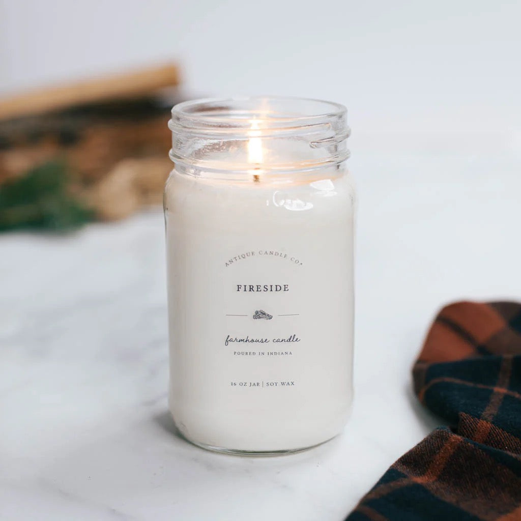 Antique Candle Co 16oz 'Fireside' soy candle lit on a marble surface with a plaid cloth. Cozy, smoky scent with long-lasting burn time.