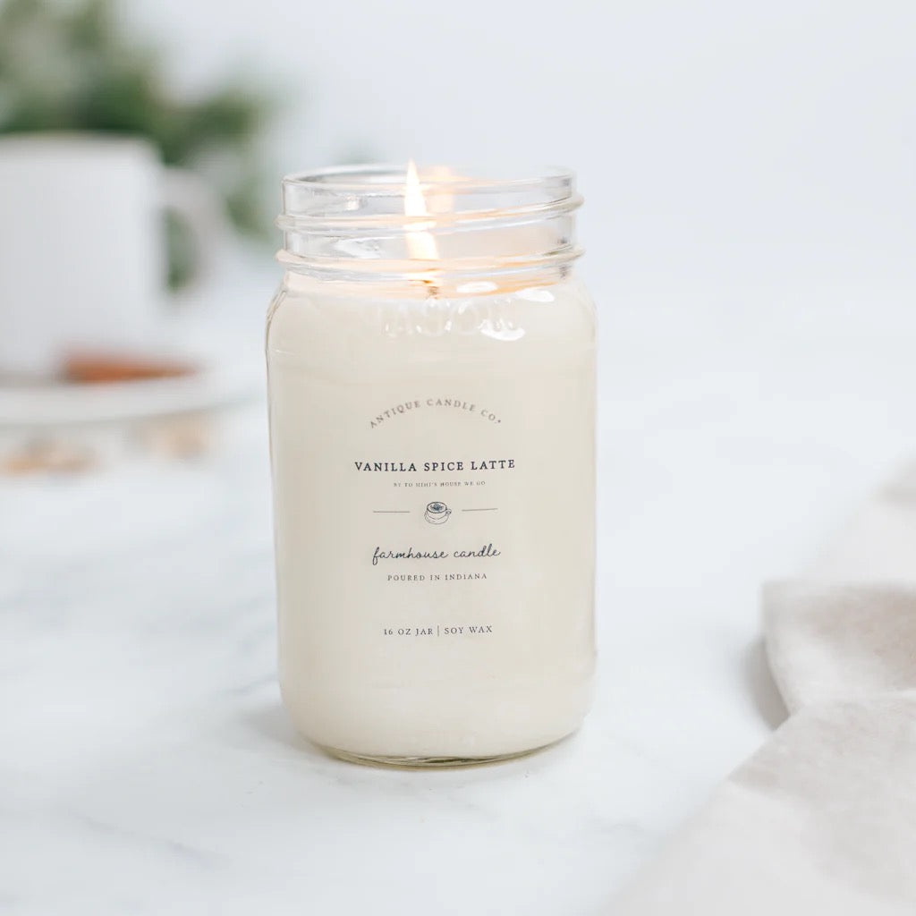 Antique Candle Co 16oz 'Vanilla Spice Latte' soy candle burning beside a coffee cup. Cozy, sweet fragrance with natural soy wax and long burn time.