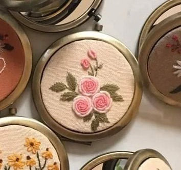 Close-up of a floral embroidered compact mirror from Quince Fables, featuring pink roses on a beige fabric cover.